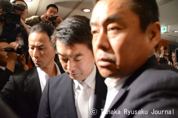警察のSPに両脇を固められ記者会見場に入る橋下大阪市長。うつ向き険しい表情だった。法廷に引きずり出されるような気分なのだろうか。=27日、正午　写真：田中龍作=